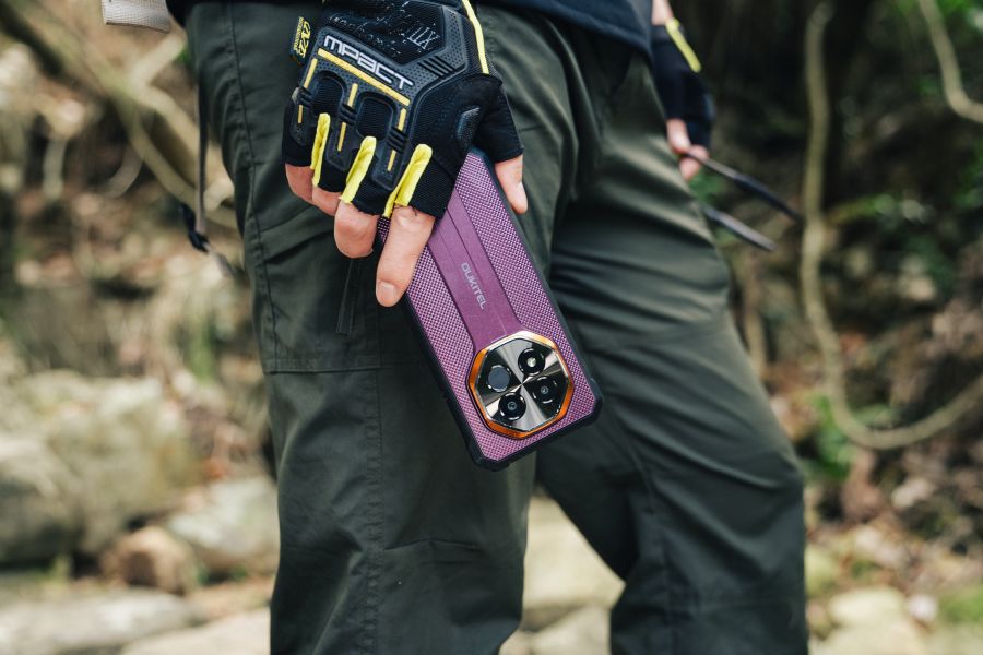 Person wearing outdoor gloves holding a purple rugged smartphone beside their leg.