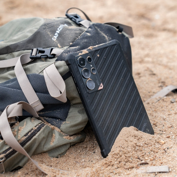 A black protective phone case with a ribbed back and camera cutouts, shown resting on a green and gray backpack in sandy terrain.