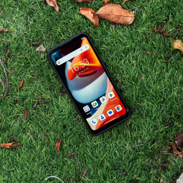 A black Oukitel WP62 rugged phone lies on green grass with scattered dry leaves. The screen is on, displaying a colorful home screen interface.